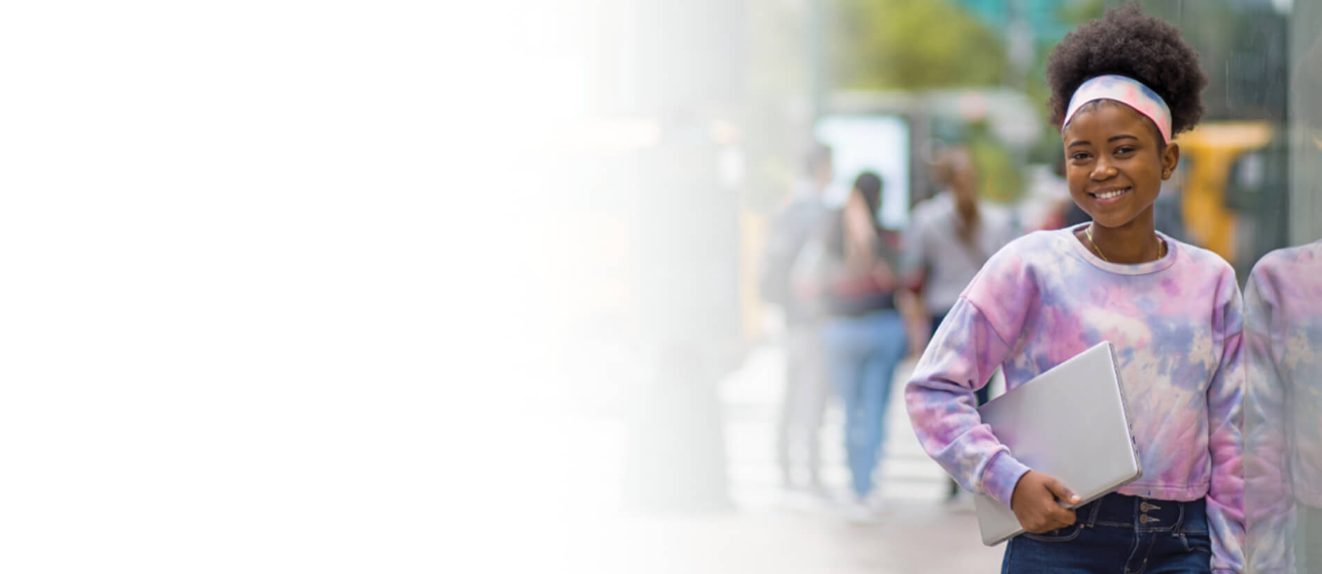 Smiling Black teenage girl in a pink tie-dye sweatshirt and headband carries a closed laptop outdoors, conveying confidence and optimism