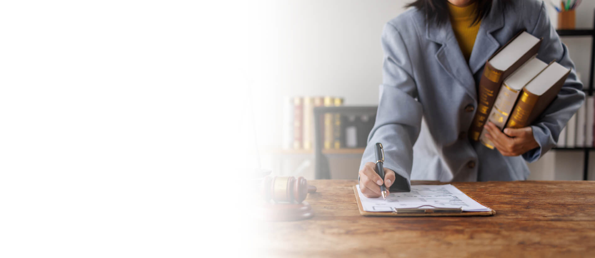 Lawyer in a gray suit writes at a wooden desk while holding law books; scales of justice and a gavel convey a serious legal setting