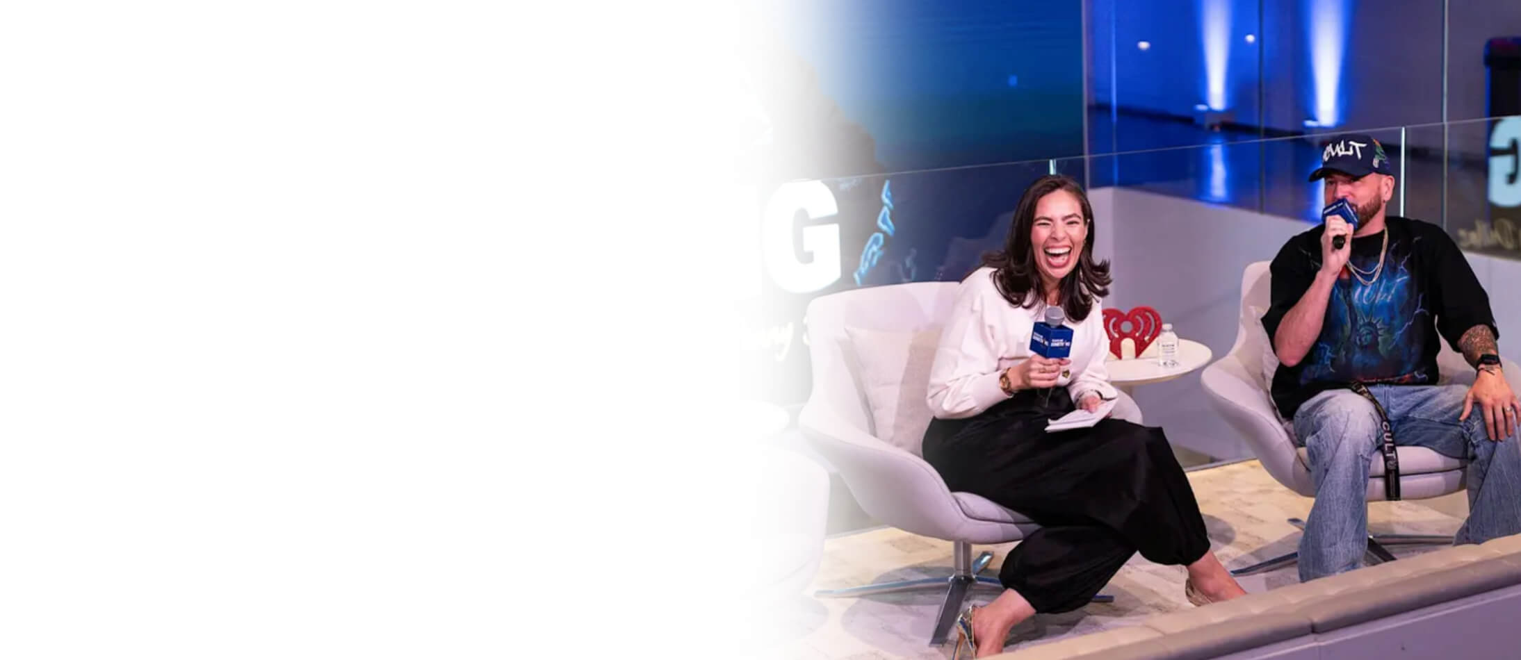 A female and male host speaking into iHeartRadio microphones during a live panel discussion on a modern stage set