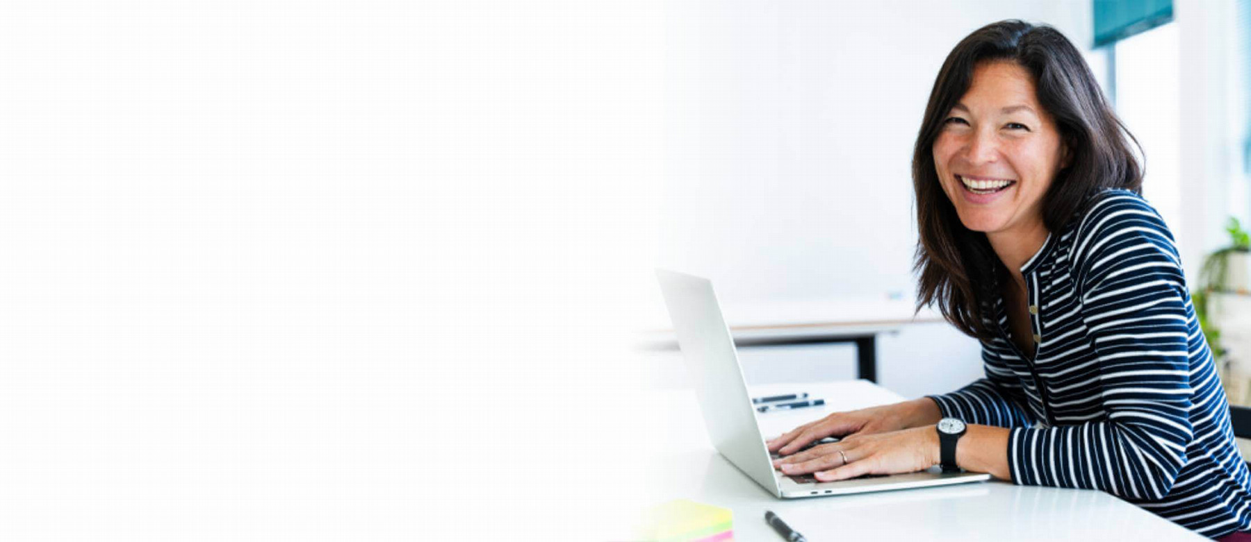 Smiling woman with long hair in a striped shirt sits at a desk, hands resting on a laptop, conveying a cheerful and approachable demeanor