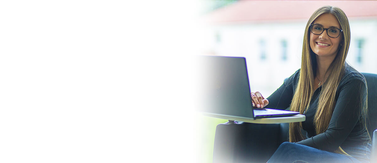 A woman with long hair sits at a table, using a laptop, in a bright, modern workspace. Sunlight filters through a nearby window.