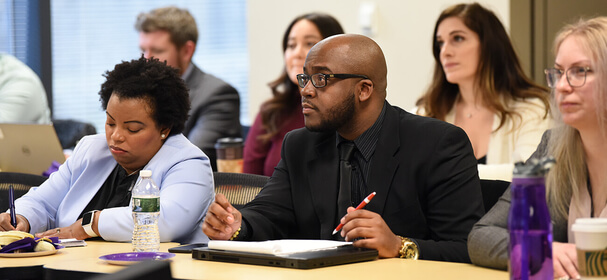Diverse professionals sit at a conference table, taking notes during a meeting; colleagues listen in the bright room, creating an attentive mood