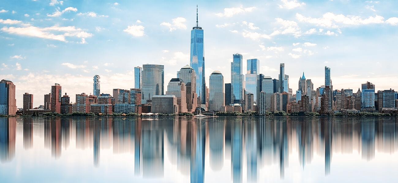 Skyline of a modern city with tall skyscrapers and mirrored reflections in the calm water below. The sky is bright with scattered clouds, creating a serene ambiance.