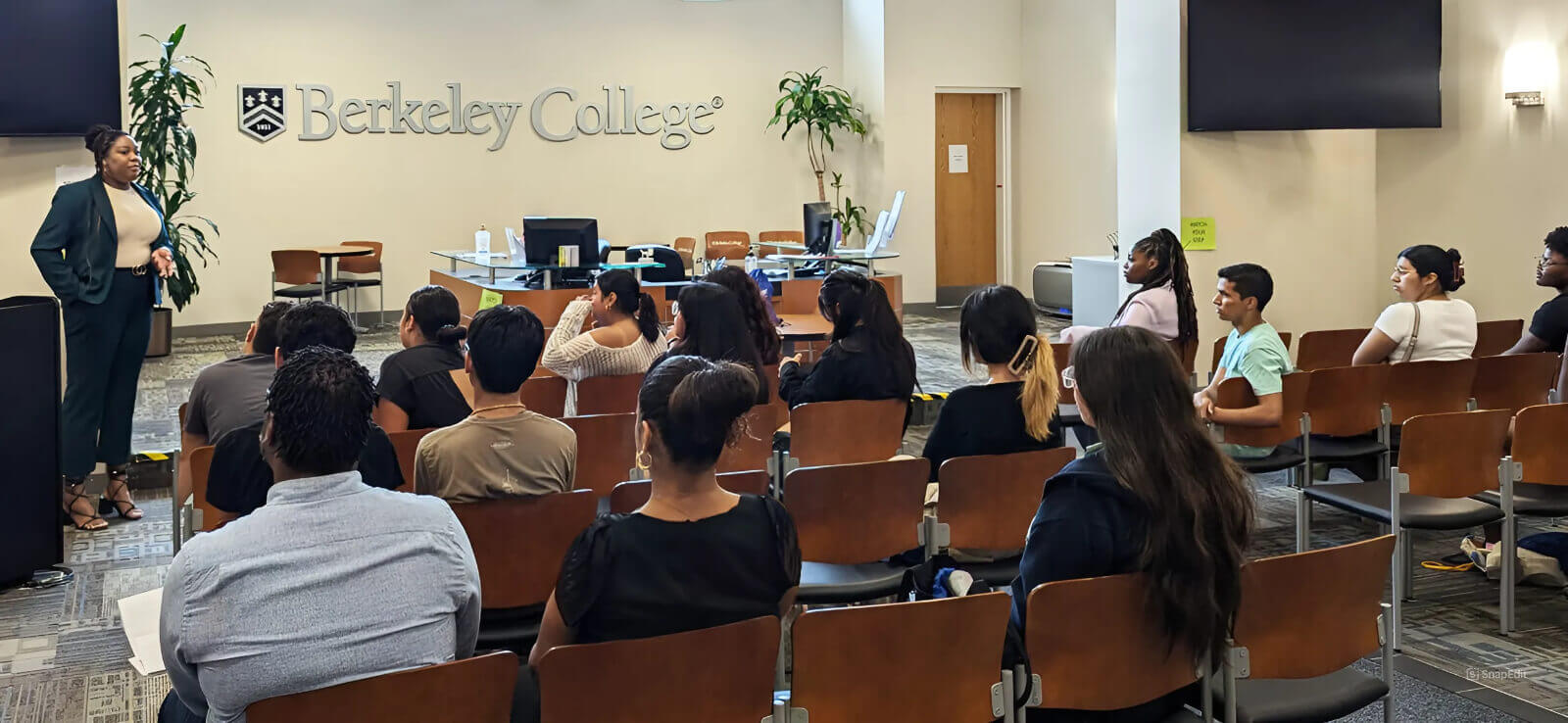 A woman stands confidently at the front, presenting to an engaged audience of diverse individuals seated in a conference room