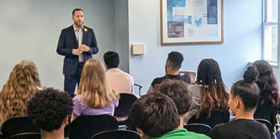 A man in a suit presents to a group of high school students, effectively communicating his presentation to the engaged audience.