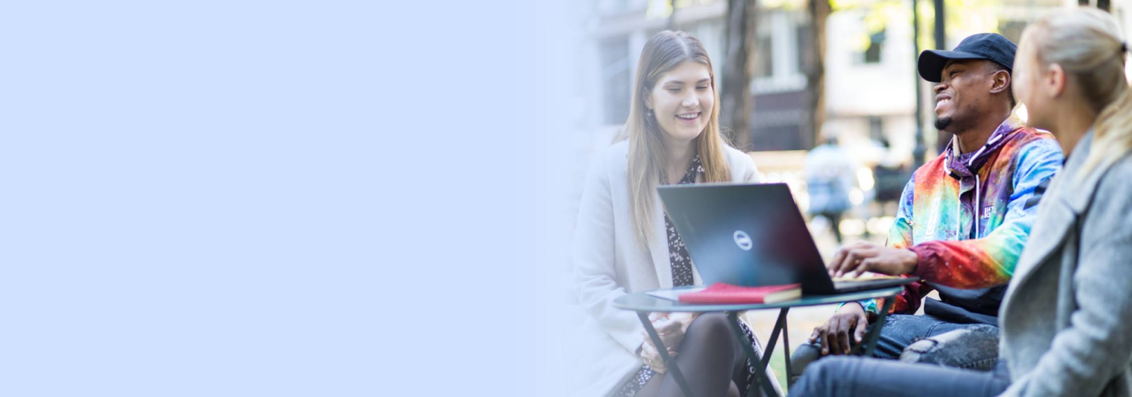 Image: A group of young people smiling and in a brightly outdoors table, there is a laptop and some books over the table