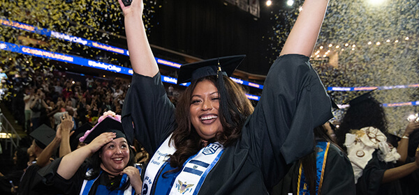 A woman in a graduation gown joyfully raises her hands in celebration