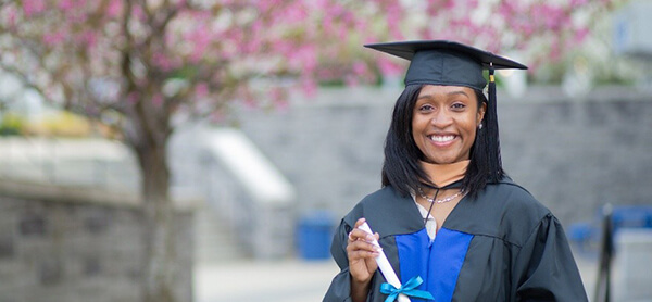 A smiling graduate in cap and gown holds a diploma. She stands in front of a blooming tree and stone building, conveying a sense of achievement and joy.