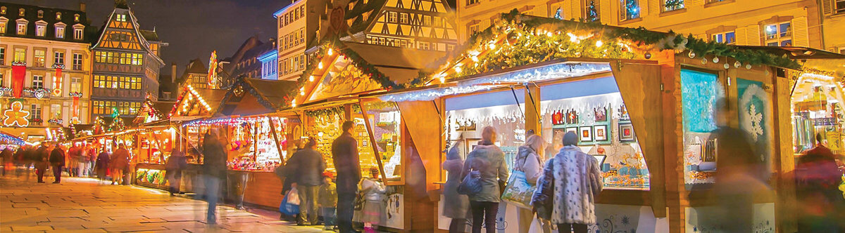 People browse illuminated wooden stalls at an outdoor Christmas market in a European town square at night, with festive decorations and historic buildings in the background.