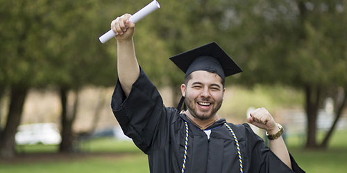 A man in a graduation gown proudly holds up his diploma, celebrating his academic achievement with a smile