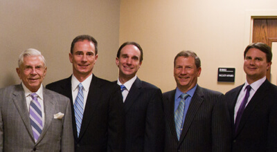 Five men in suits, standing side by side against a plain wall. They have blurred faces and are in a professional setting.