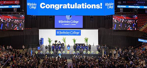 Graduation ceremony at Berkeley College with graduates on stage and a large blue: Congratulations, banner overhead. Audience seated, celebrating.