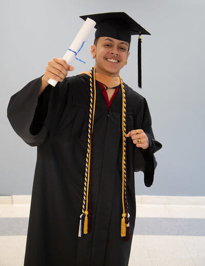 Photo of Jianni Enzo de Jesus. Young man in graduation attire, smiling and raising his arm triumphantly, holding a diploma. His expression conveys joy and accomplishment.