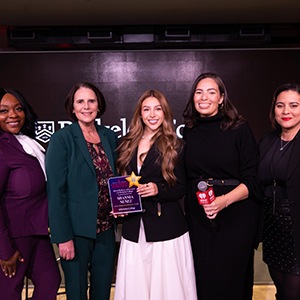 Five diverse women stand in a row, and the woman in the center holds an award.