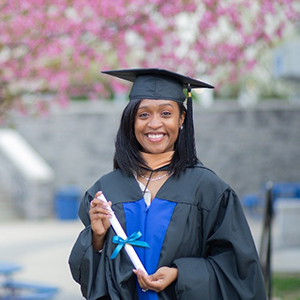 A woman smiles in a cap and gown in front of cherry blossom trees.