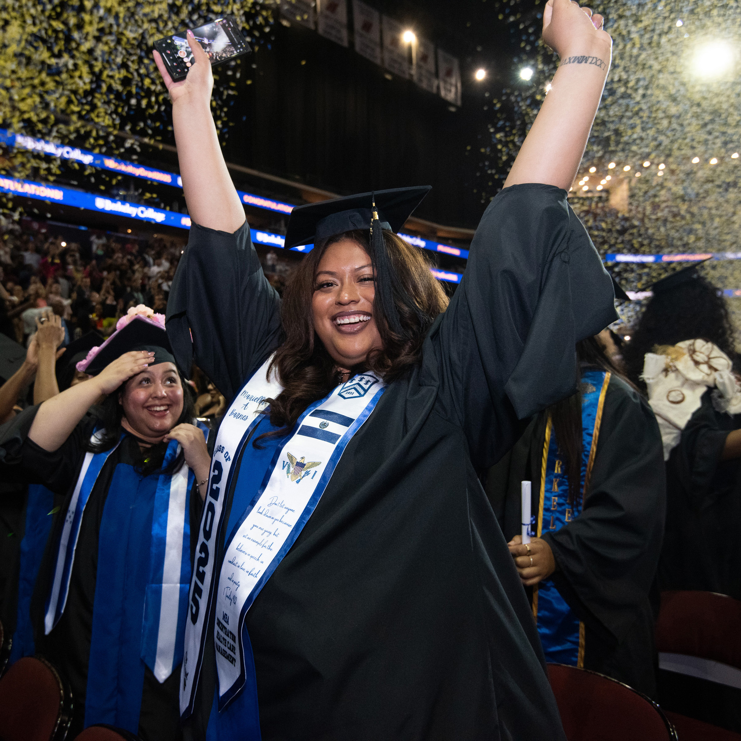 A woman in graduation regalia raises her hands amid confetti