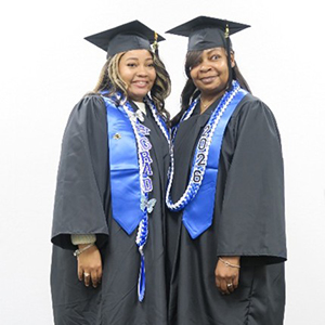 A mother and daughter smile in graduation caps and gowns.