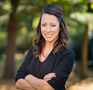 A woman with crossed arms poses confidently in front of a backdrop of vibrant trees