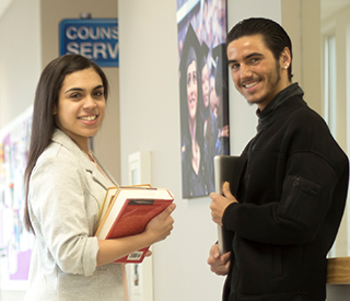 Photo of 2 students holding books in hallways with 3 students in the background mobile image
