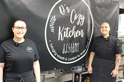 Two women in aprons stand beside a sign that reads "D's Cozy Kitchen,"