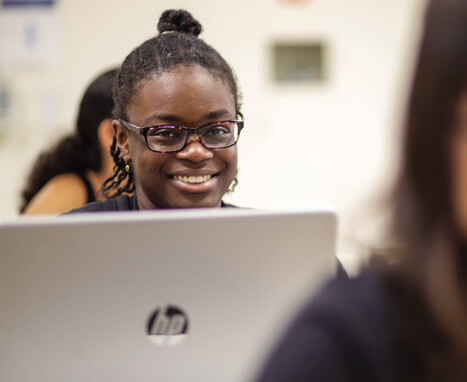 A student sits with a laptop open in a classroom, focusing on their work while others are visible in the background.
