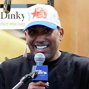 A person smiles widely, wearing a cap, holding a microphone with a blue cover, standing in front of a sign. The atmosphere is lively.