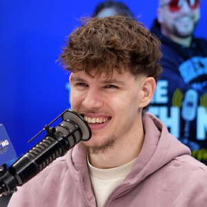 Photo of Liam Winn, Young man with curly hair and a goatee smiles while speaking into a microphone. He wears a pink hoodie. Background shows a blue poster with text.
