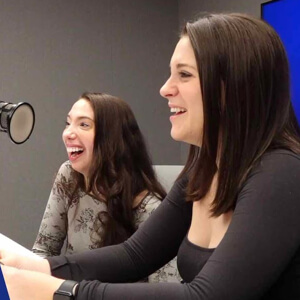 Two women seated at a table, engaged in a discussion. A microphone is visible in the foreground, with a modern backdrop.