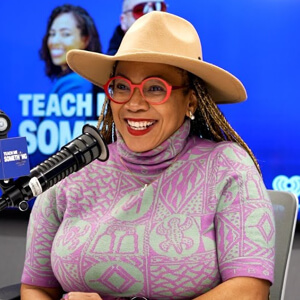 A person wearing a stylish wide-brimmed hat and a patterned purple sweater sits at a microphone in a modern studio setting.