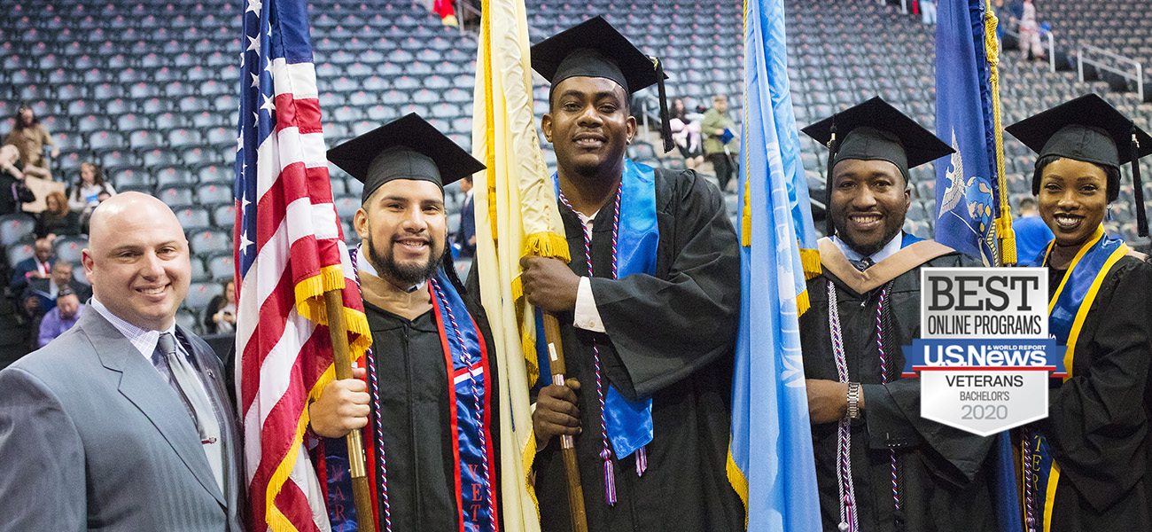 Group of 3 male and 1 female military students at Commencement in cap and gown holding flags with male and female