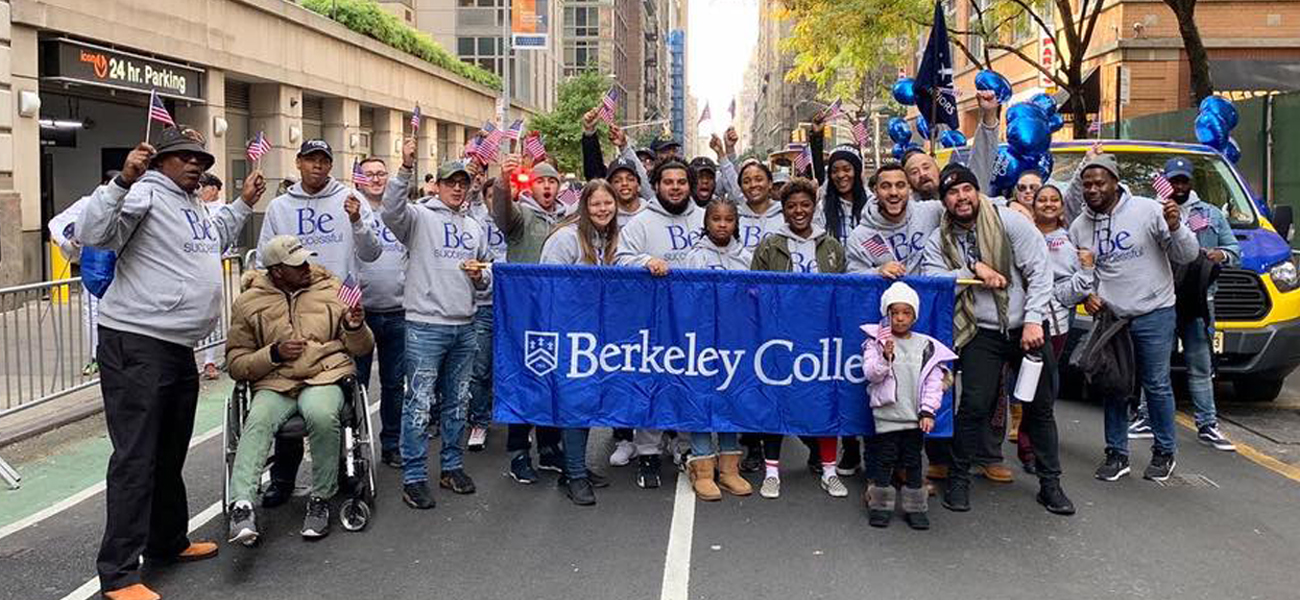 Large group of Berkeley College Military and Veterans students and staff at the Veterans Day Parade holder Berkeley College banner and wearing gray Be Successful sweatshirts 