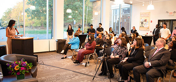 A woman stands speaking at a podium to an attentive audience seated indoors. The room features large windows, flowers, and a relaxed, engaged atmosphere.