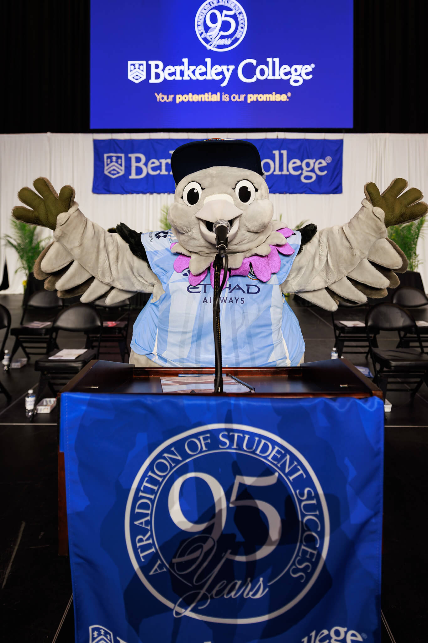 A pigeon mascot stands at a podium with a blue 95th anniversary banner.
