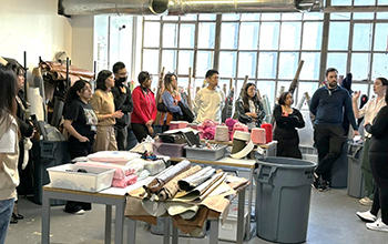 Students stand in a warehouse and in the center is a table covered in rolled textiles and fabrics.
