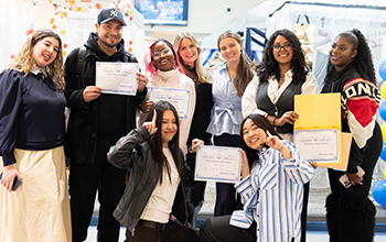 A group of diverse students smiles and hold certificates awarding them money for various cotton-based designs they created for a competition.