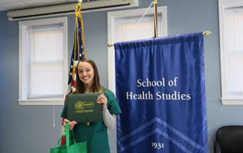 A woman in scrubs holds an award standing in front of two flags.