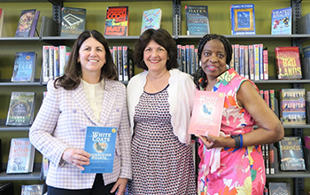 A group of three diverse women stand in front of a bookshelf smiling.