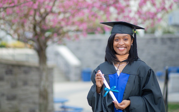 A woman in graduation regalia stands smiling in front of cherry blossom trees.