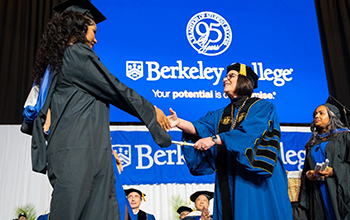 A Black woman in graduation regalia receives her diploma from a female college president dressed in doctoral graduation regalia.