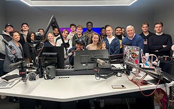 Students, associate, and radio hosts smile for a photo amidst radio technology including microphones, laptops, and broadcasting equipment.