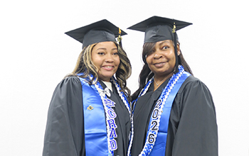 A mother and daughter stand smiling side by side in graduation regalia.