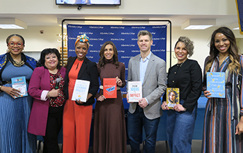Seven diverse people stand in a row, six holding books they authored.