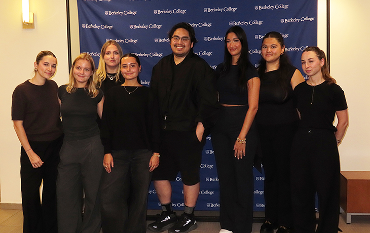 Group of eight diverse students standing and smiling in front of a Berkeley College backdrop indoors.