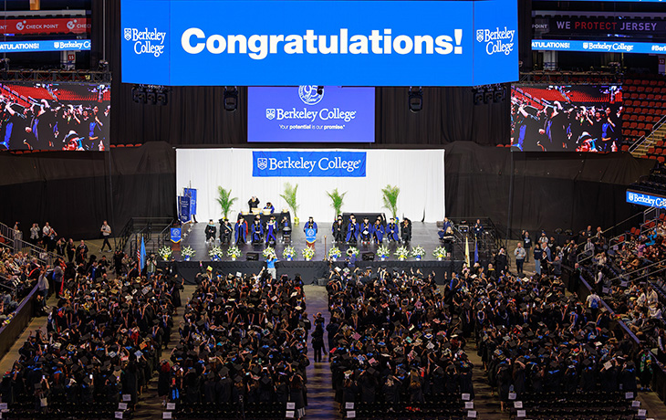 Graduation ceremony at Berkeley College with graduates in caps and gowns, faculty on stage, and a large "Congratulations!" sign overhead.