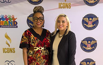 Two women stand in front of a backdrop that says The Hand Foundation with the presidential seal
