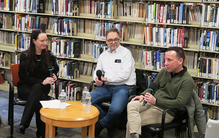 a woman and two men with microphones sit at a table in front of a shelf of books