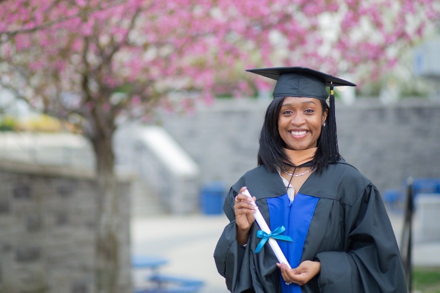 A woman in graduation regalia stands and smiles in front of cherry blossom trees.