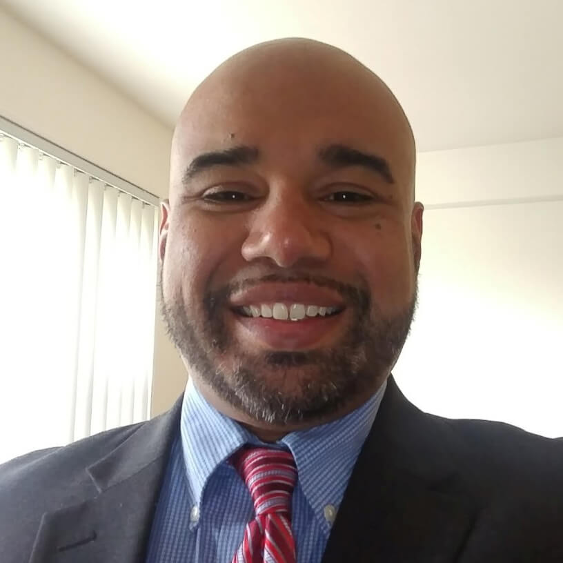 Smiling man with a trimmed beard wearing a dark suit, blue shirt, and red striped tie in a bright room with vertical blinds.