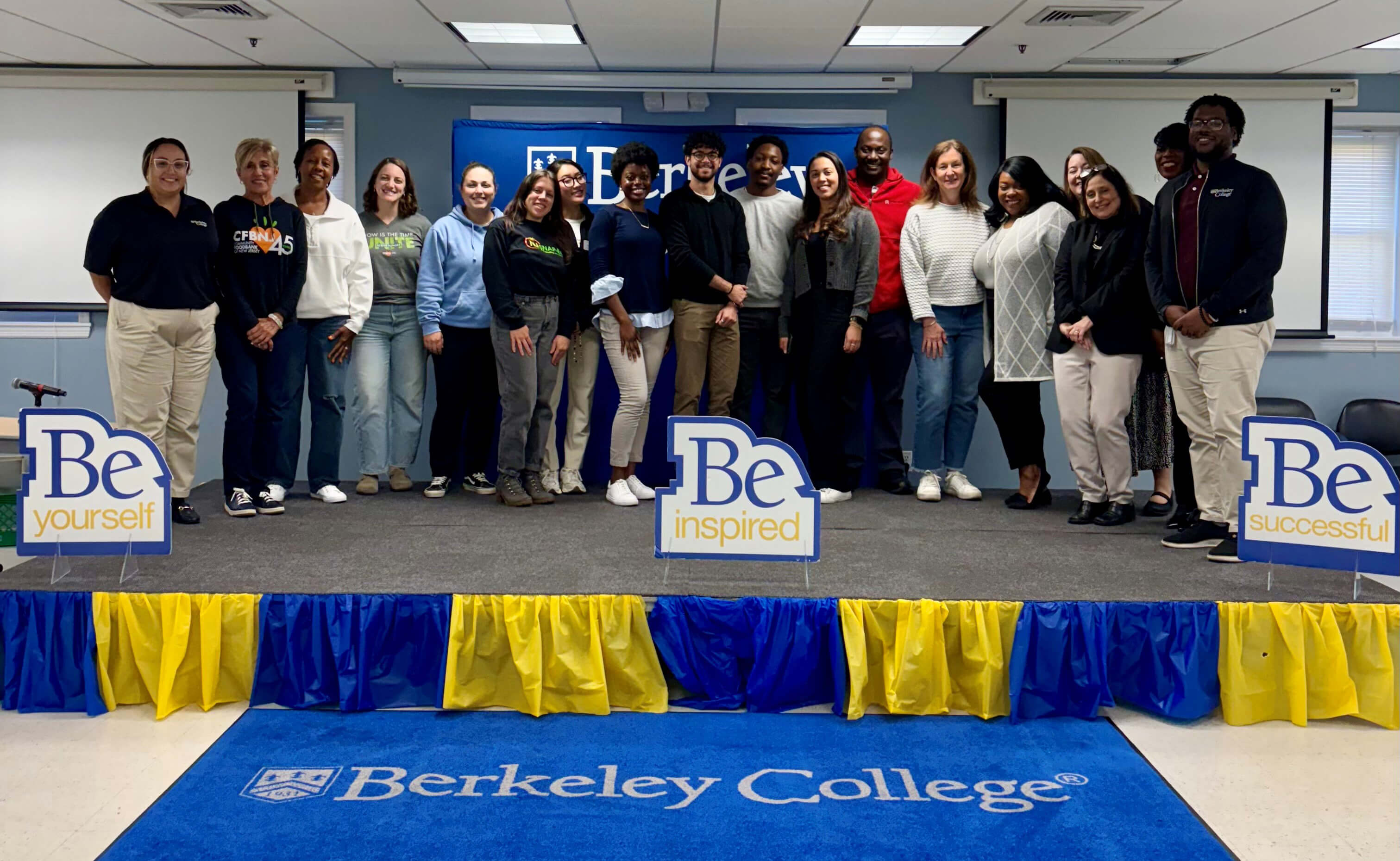 Group of diverse students standing on a stage with Berkeley College signs reading "Be yourself," "Be inspired," and "Be successful."
