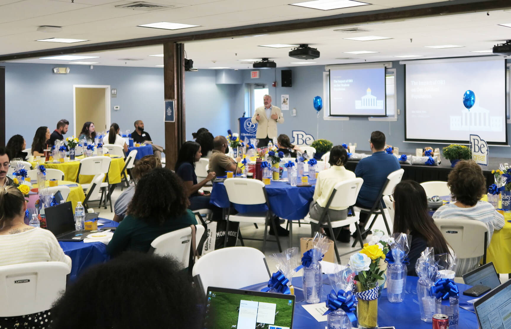Speaker presents to an audience at a decorated banquet event with blue-and-yellow tables, gift baskets, and projection screens.